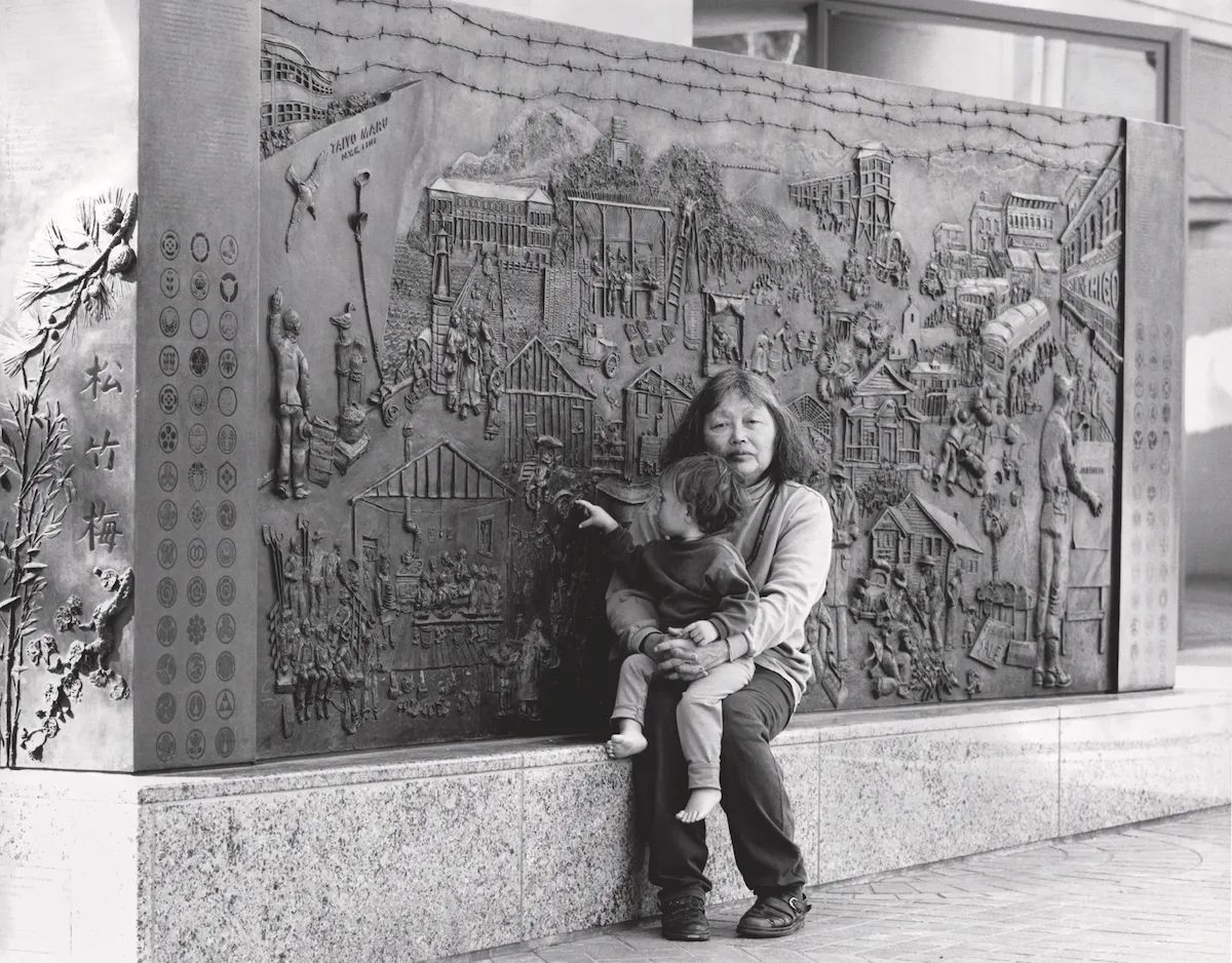 日系アメリカ人収容記念碑《Japanese American Internment Memorial (PC.011)》(1990–94)の前に孫娘と座るルース・アサワ。Photo: ©2025 Ruth Asawa Lanier, Inc./Courtesy David Zwirner/Photo Laurence Cuneo