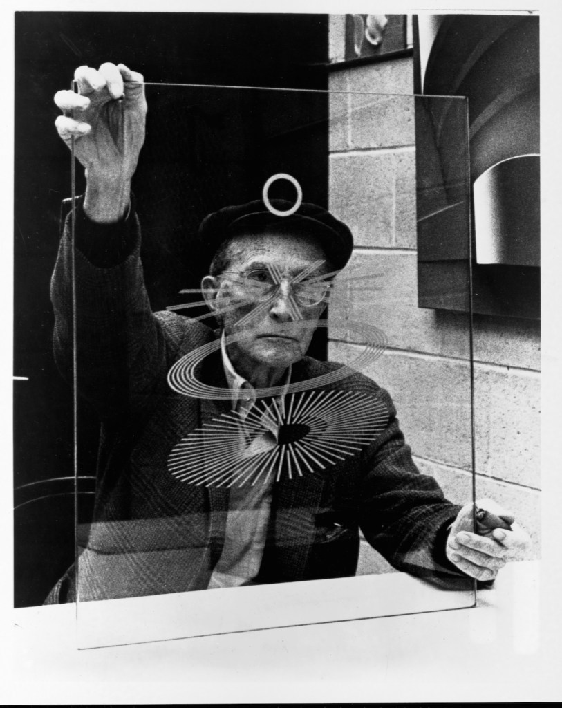 リチャード・ハミルトンがデュシャンと共同制作した《Oculist Witnesses》（1968）を持つマルセル・デュシャン。これは、ハミルトンが1966年に再構築したデュシャンの《彼女の独身者たちによって裸にされた花嫁、さえも（大ガラス）》（1915-1923）と関連するマルチプル作品。　Photo: Library of Congress/Corbis/VCG via Getty Images